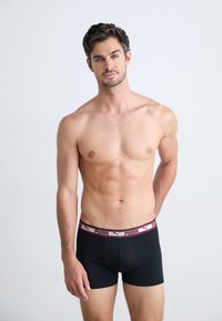 Felsőtest nélküli férfi, rövid, sötét hajjal, fekete Puma boxer alsót visel, egy sima világos háttér előtt állva, a kamerába néz.