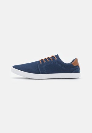Marineblaue Freizeitsneaker mit weißer Sohle, braunen Schnürsenkeln und braunem Fersenakzent, von der Seite auf weißem Hintergrund gezeigt.