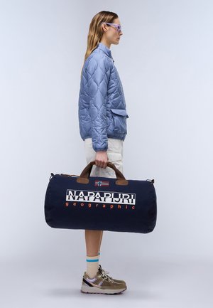 Borsa da viaggio navy con manici marroni, caratterizzata da un logo bianco e arancione. Indossata con una giacca trapuntata azzurro chiaro e pantaloni beige.