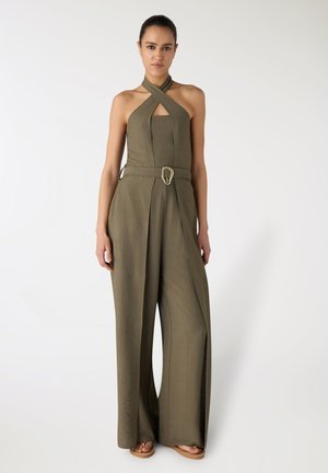 Donna che indossa una tuta verde oliva con scollo halter, gambe larghe e cintura con fibbia dorata, in piedi su sfondo bianco.
