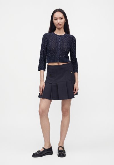Femme portant un cardigan court en maille foncé, une mini-jupe plissée avec poches à boutons, et des chaussures Mary Jane noires, debout devant un fond uni.