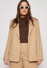 Beige blazer med slagna kragar och fickor, bärs över en brun topp. Modellen har även på sig vita kantiga solglasögon.