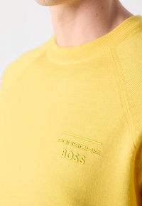 Gros plan sur une personne portant un pull jaune avec les logos "Porsche" et "BOSS" en relief sur la poitrine.
