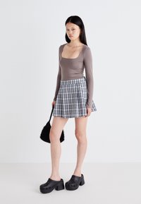 Graues Langarmoberteil, karierte, gefaltete Minirock in Schwarz und Weiß. Schwarze Plateausandalen und eine strukturierte schwarze Schultertasche runden das Outfit ab.