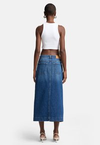 Jupe maxi en denim bleu moyen avec une coupe droite, dotée de deux poches arrière et d'une étiquette en cuir sur la taille. Associée à un débardeur court blanc.