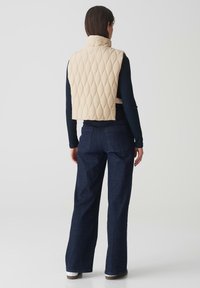Beige gesteppte Weste mit hohem Kragen, cropped Design, kombiniert mit dunkelblauen Schlagjeans und weißen Sneakers. Glatte Textur, minimale Akzente.