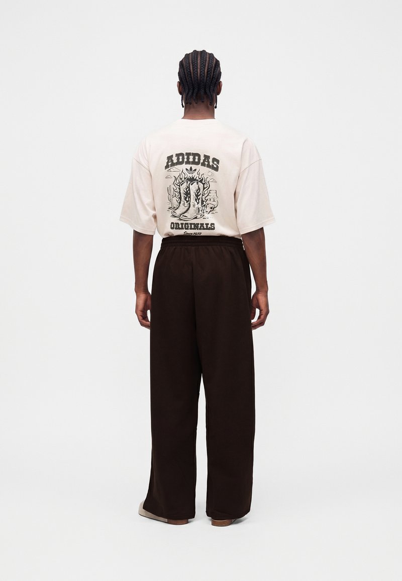 Person med fletninger iført en beige Adidas Originals t-shirt med støvlegrafik og sorte vidde bukser, der står med ryggen mod på en hvid baggrund.