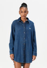 Denim krekla kleita, tumši zila, ar pogu priekšpusē, norādītu apkakli, garām piedurknēm, ar nelielu plāksteri uz kreisās krūts puses, taisna griezuma ar sānu slīpēm.