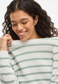 Femme souriante aux cheveux bouclés tenant le col d'un pull en tricot blanc et vert à rayures, regardant vers le bas.