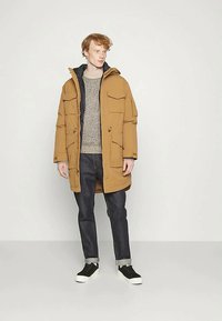 Νέος άνδρας που στέκεται φορώντας μια καφέ μπεζ καπιτονέ ζακέτα με κουκούλα, γκρι πουλόβερ, σκούρα τζιν αναδιπλωμένα και μαύρα αθλητικά παπούτσια σε απλό ανοιχτό φόντο.