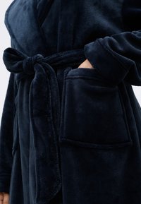 Laiva sininen, pehmeä kylpytakki, jossa on liivikaulus, sidottava vyö ja sivutaskut. Kankaan pinta on pehmeä ja teksturoitu.