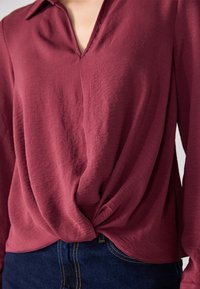 Burgunderfarget langermet skjorte med V-hals, med et design med knute foran og myk, teksturert stoff. Kombinert med mørkeblå jeans.