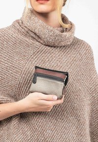 Mujer con un suéter de punto beige sosteniendo cerca de su pecho un pequeño monedero de cuero marrón y beige con cremallera.