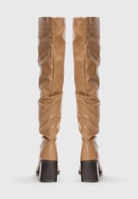 Bottes en cuir beige hautes avec des tiges molles et de larges talons blocs brun foncé, vues de l'arrière sur un fond blanc.