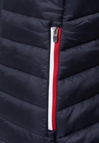 Marinblå quiltad pufferjacka med en dragkedjeficka och röd- och vitakenter längs dragkedjan. Slät, lätt tygtextur.