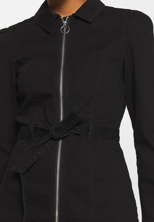 Czarna sukienka na długi rękaw z kołnierzykiem, srebrnym przednim zamkiem błyskawicznym z kółkiem do ściągania oraz wiązanym węzłem z materiału w talii na stojącej osobie.