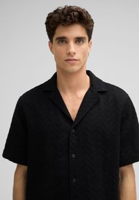 Schwarzes Kurzarmhemd mit einem strukturierten Zickzackmuster, einem eingekerbten Kragen und fünf Knöpfen vorne zur Schließung.