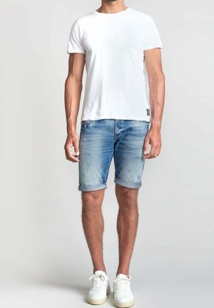 Mand iført en ensfarvet hvid kortærmet t-shirt, falmede blå op rullede denimshorts og hvide sneakers, stående på en grå baggrund.