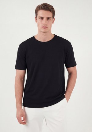Jeune homme aux cheveux courts et bruns portant un t-shirt noir uni à manches courtes et un pantalon blanc, debout avec une main dans la poche.