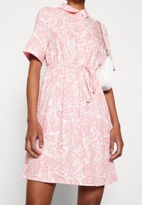 Vestido de manga corta y cuello en rosa claro con un patrón de paisley. Presenta una cintura ajustada y una falda fluida. Se ve un bolso blanco.