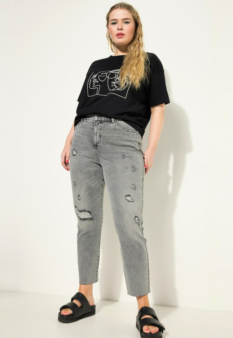 Camiseta negra con un gráfico lineal de una cara, combinada con jeans rectos desgastados de color gris claro y sandalias negras con tiras gruesas.