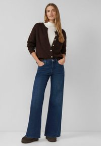 Vrouw staat met handen in haar zakken, draagt een donkerbruine gebuttonde cardigan, een witte coltrui, wijde blauwe jeans en donkere schoenen, op een grijze achtergrond.