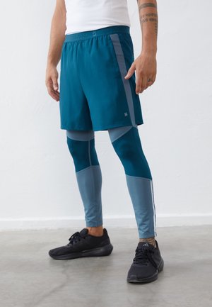 Homme portant un short bleu sarcelle par-dessus des leggings de compression bicolores bleus et des baskets noires, debout sur un sol gris contre un mur blanc.