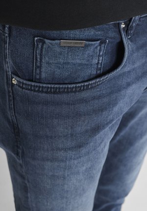 Nahaufnahme der Tasche einer blauen Jeans mit sichtbaren Nieten und einem kleinen schwarzen Etikett mit der Aufschrift "ANTONY MORATO" über der Münztasche.