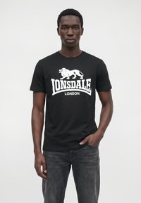 Tânăr stă cu fața înainte purtând un tricou negru Lonsdale London și blugi spălați închis, cu o mână în buzunar.