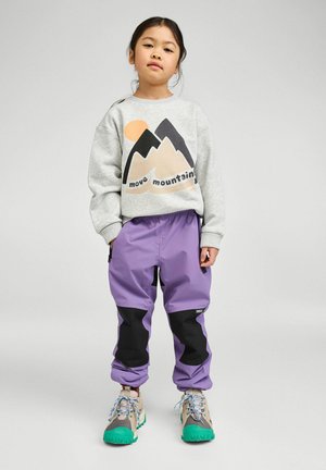 Młoda dziewczyna stojąca, ubrana w szarą bluzę z grafiką gór i napisem „move mountains”, fioletowe spodnie oraz buty trekkingowe z zielonymi akcentami.