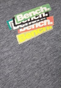 Camiseta gris con logos en capas de "Bench." en verde, negro, rojo y amarillo, con una tela texturizada y un diseño informal.