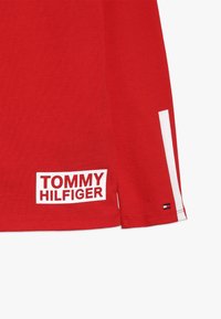 Červená bavlněná košile s bílým obdélníkovým logem s nápisem "TOMMY HILFIGER." Obsahuje bílé boční pruhy a štítek s logem.