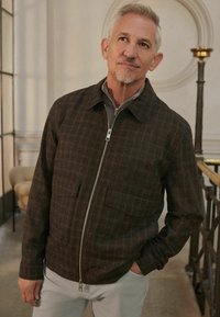Brown plaid zip-up jacket with a collared neck, featuring two front patch pockets and a smooth texture. Worn over a gray top.