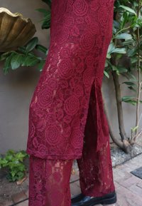 Burgundske čipkaste hlače z rožnatimi vzorci, polprosojno teksturo in stranskimi zarezi, v kombinaciji s črnimi čevlji na zelenem ozadju.