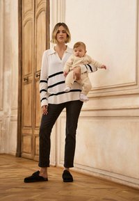 Suéter de rayas de manga larga en crema y azul marino sobre vaqueros oscuros, combinado con zapatos negros sin cordones; la modelo sostiene un conjunto beige para bebé.