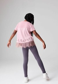 Top rosa a maniche corte con bordo in pizzo, con la scritta 'ALLSAINTS' ricamata sul retro. Abbinato a leggings grigi e scarpe da ginnastica bianche.