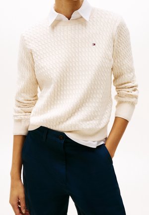 Personne portant un pull torsadé crème par-dessus une chemise blanche à col avec un pantalon bleu marine, une main dans la poche.