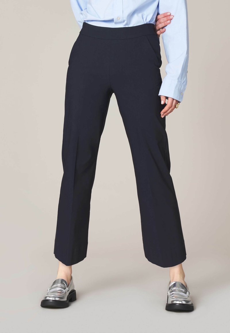 Personne portant un pantalon noir à jambes larges, une chemise bleu clair boutonnée et des mocassins argentés, debout une main sur la hanche devant un fond neutre.