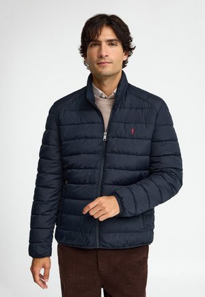Uomo con capelli scuri che indossa una giacca trapuntata blu navy e pantaloni marroni su uno sfondo bianco uniforme.