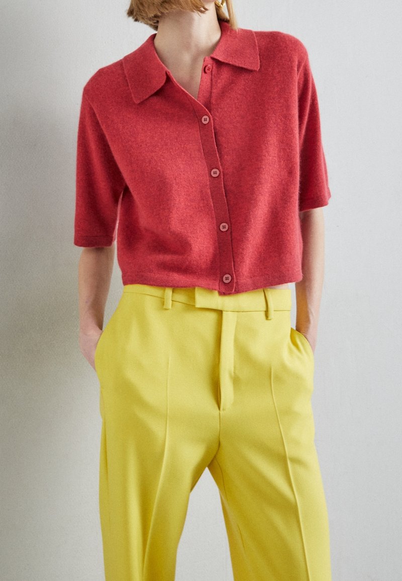 Femme portant une chemise rouge à manches courtes boutonnée, rentrée dans un pantalon jaune taille haute avec les mains dans les poches.