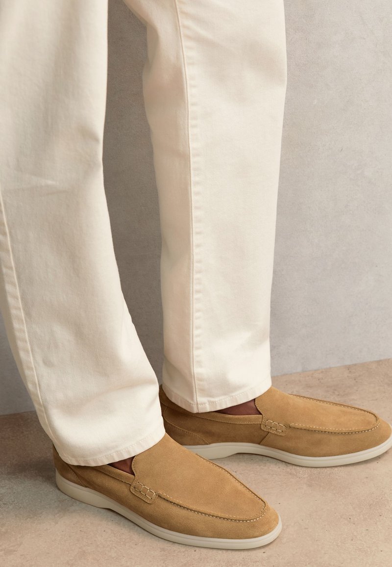 Hombre con zapatos beige de ante sin cordones con suelas blancas y pantalones color crudo, de pie sobre un suelo claro frente a una pared gris.