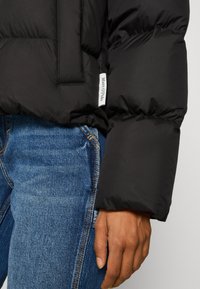 Schwarze Pufferjacke mit einem gesteppten Design, ausgestattet mit einem sichtbaren Etikett und einer glatten, glänzenden Textur. Getragen über blauen Jeans.