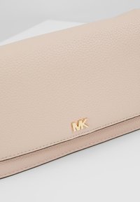 Portefeuille en cuir texturé beige avec coutures visibles et petit logo MK doré sur le rabat avant, posé sur une surface blanche.
