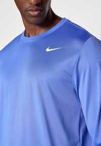 Langærmet blå sportsbluse med en glat tekstur, med et hvidt Nike-logo på venstre øverste side. Simpelt rund halsudskæring design.