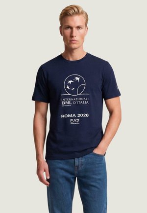 Młody mężczyzna w granatowej koszulce z białym logo ptaka i napisem "Internazionali BNL d'Italia Roma 2026", w połączeniu z niebieskimi jeansami.