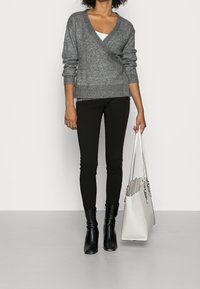 Grauer Strickpullover mit langen Ärmeln, kombiniert mit schwarzen Hosen und eleganten schwarzen Knöchelstiefeln. Hält eine große weiße Tragetasche.