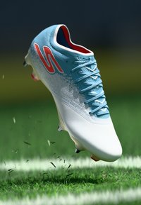 Crampon de football bleu clair avec des accents blancs, tige texturée et lacets, flottant au-dessus de l'herbe, montrant des crampons rouges et noirs.