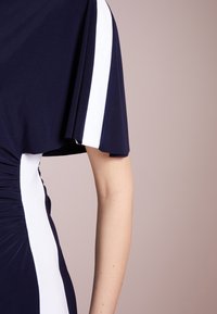 Marinblå klänning med vita sidostripes, tillverkad av mjukt material, med lösa ärmar och rynkad struktur längs midjan. Fokuserar på överkroppen.