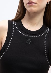 Κοντινό πλάνο γυναίκας που φοράει μαύρο αμάνικο τοπ με ασημένιες τρουκς που τονίζουν το ντεκολτέ και τους ώμους, με το λογότυπο DKNY κεντημένο στο στήθος.