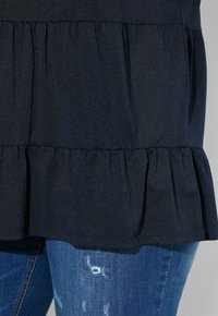 Černý texturovaný top s vrstveným volánkovým lemem. V kombinaci s modrými džínami s distressed vzorem.